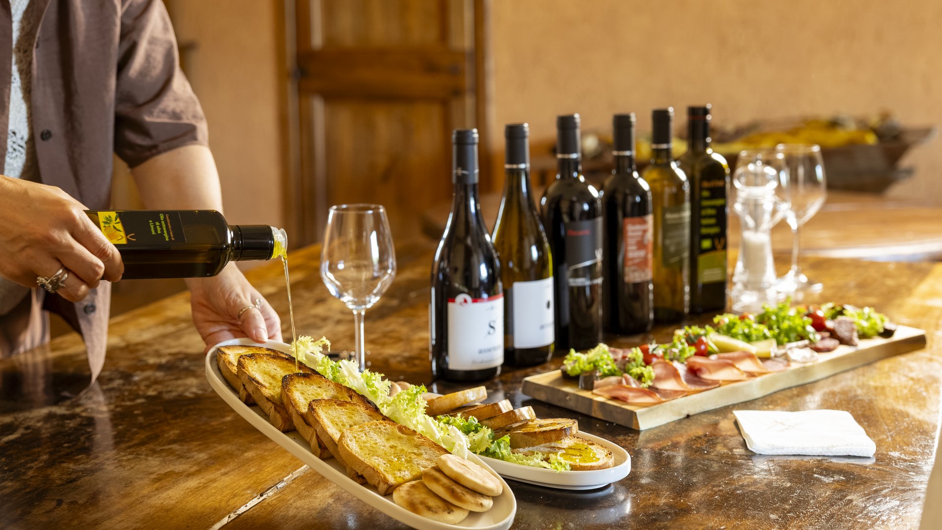 Pranzo di Pasquetta ad Assisi con Percorso SPA A Valle di Assisi, il Ristorante Recanto vi aspetta Persona versa olio d'oliva su pane tostato con bottiglie di vino e antipasti sul tavolo